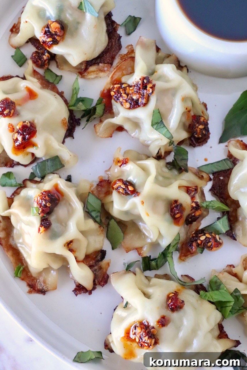 Thai Basil Chicken Dumplings Recipe with Chili Soy Dipping Sauce 12 cooked blanket dumplings on a plate topped with thai basil and chili crisp, with a small bowl of dipping sauce on the side