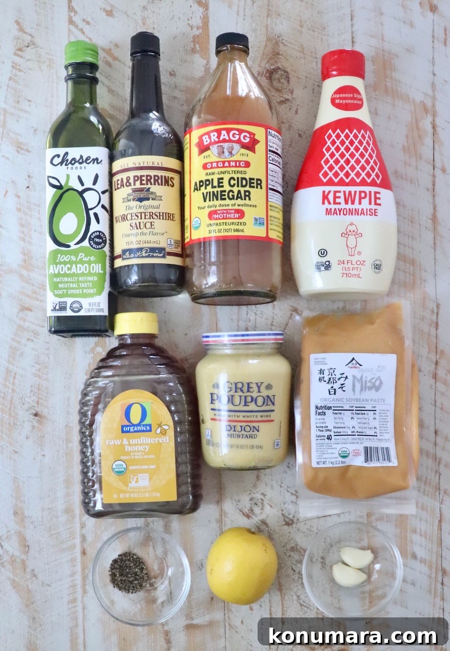 A variety of ingredients are arranged on a wooden surface, including avocado oil, Worcestershire sauce, apple cider vinegar, Kewpie mayonnaise, honey, Dijon mustard, miso paste, a lemon, garlic cloves, and a small bowl of pepper.