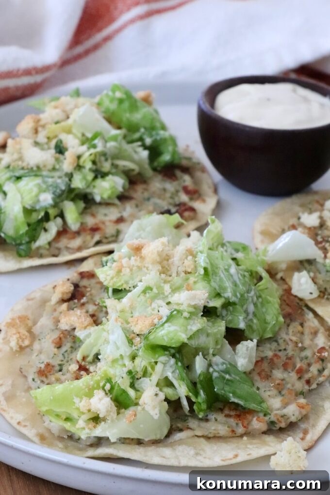 Two smash chicken tacos topped with leafy green salad and crumbled croutons on a white plate. A small bowl of creamy dipping sauce is on the side.