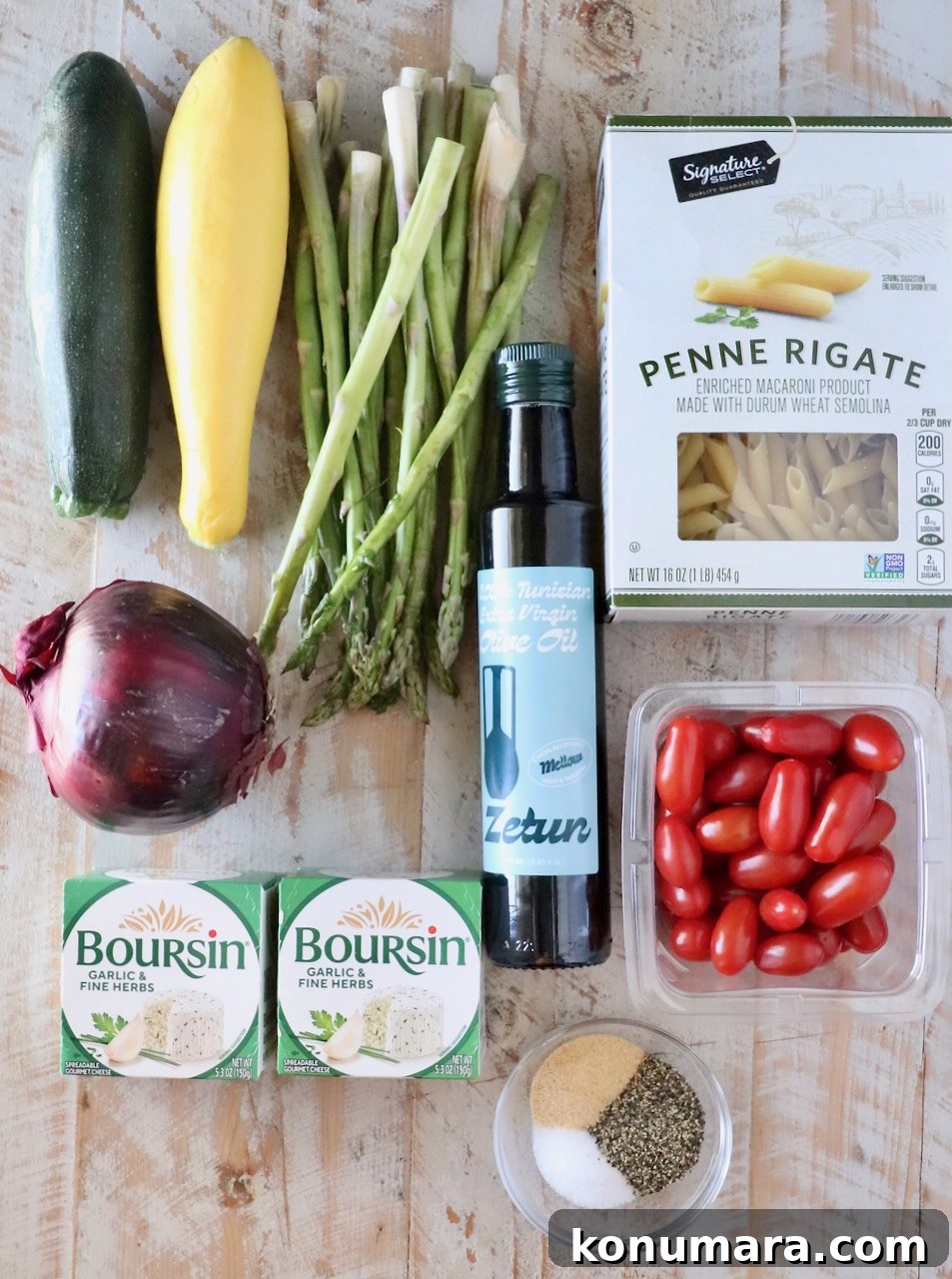 Assorted fresh ingredients on a wooden surface: zucchini, yellow squash, asparagus, red onion, cherry tomatoes, penne pasta, olive oil, Boursin cheese, and spices.