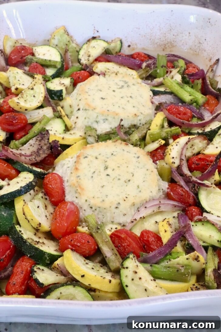 Roasted vegetables with two rounds of baked boursin cheese in a white baking dish. The veggies include zucchini, cherry tomatoes, asparagus, and red onions, seasoned with salt and pepper.