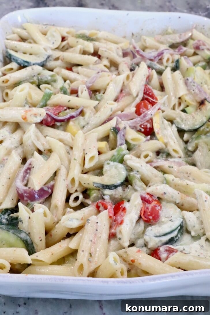 Creamy pasta bake with penne, zucchini, cherry tomatoes, red onions, and asparagus in a white casserole dish.