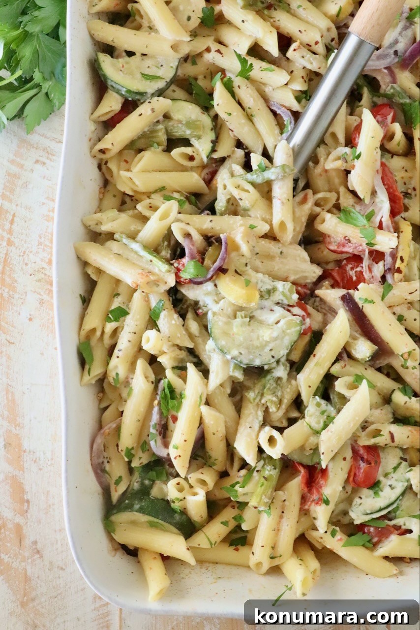 Creamy penne pasta in a white dish with zucchini, red onion, and tomatoes. Garnished with parsley.