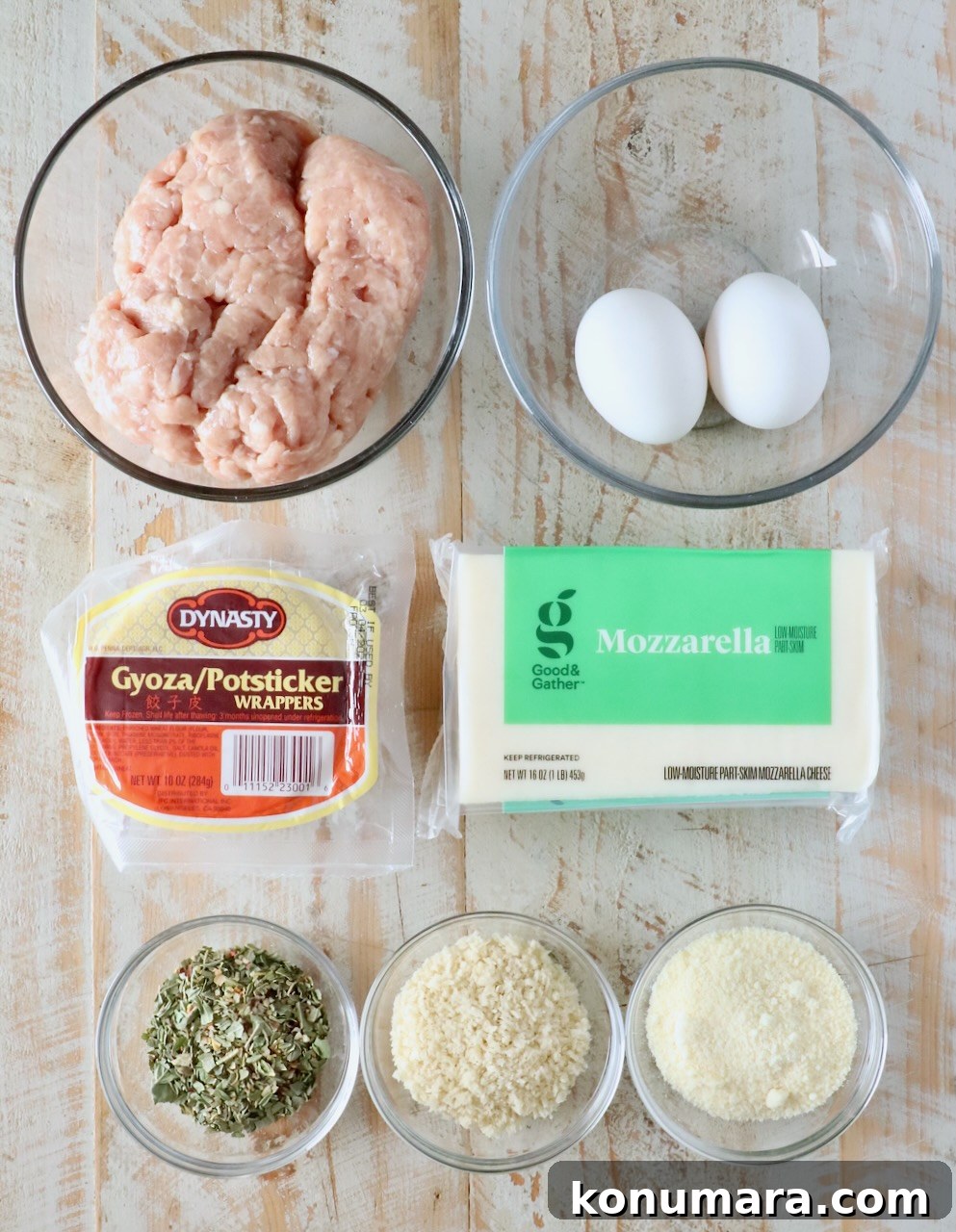 A top-down view of cooking ingredients on a wooden surface: ground chicken, two eggs, gyoza wrappers, mozzarella cheese, herbs, breadcrumbs, and grated cheese in bowls.