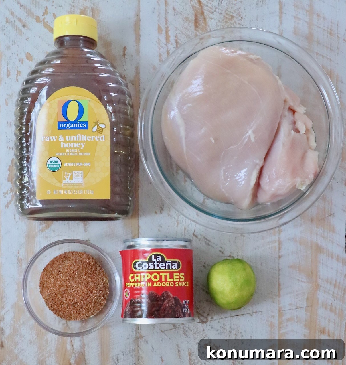 Ingredients for cooking are arranged on a wooden surface: raw honey in a bottle, raw chicken breasts in a bowl, chipotle peppers in a can, a lime, and a bowl of taco seasoning.