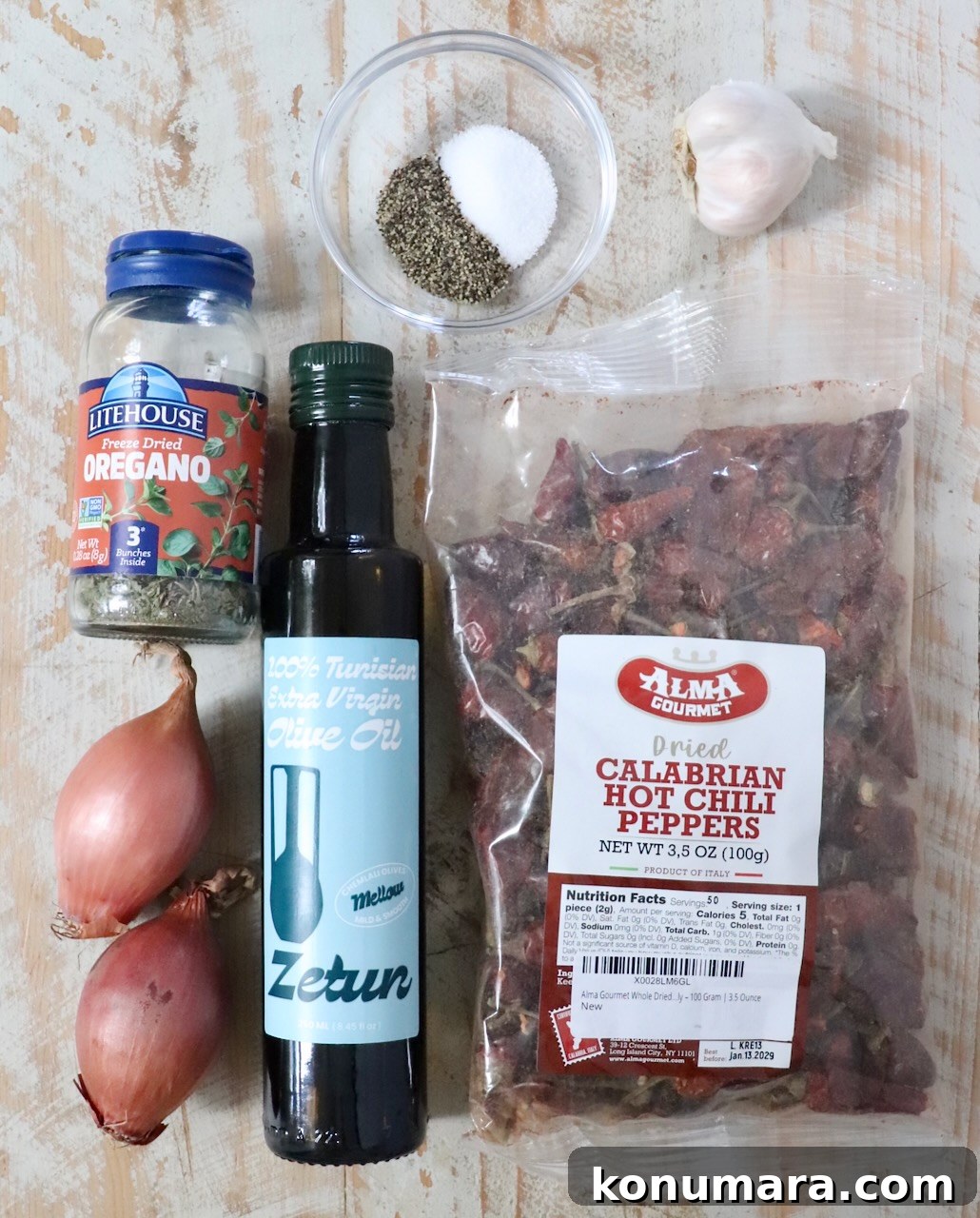Ingredients on a rustic wooden surface include a bottle of olive oil, a jar of dried oregano, dried Calabrian chili peppers, two shallots, a garlic bulb, and a bowl of salt and pepper.