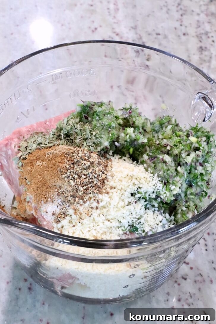 Gyro-Style Meatballs with Tzatziki Sauce 8 Glass bowl with minced meat, breadcrumbs, chopped herbs, and spices on a marble countertop.