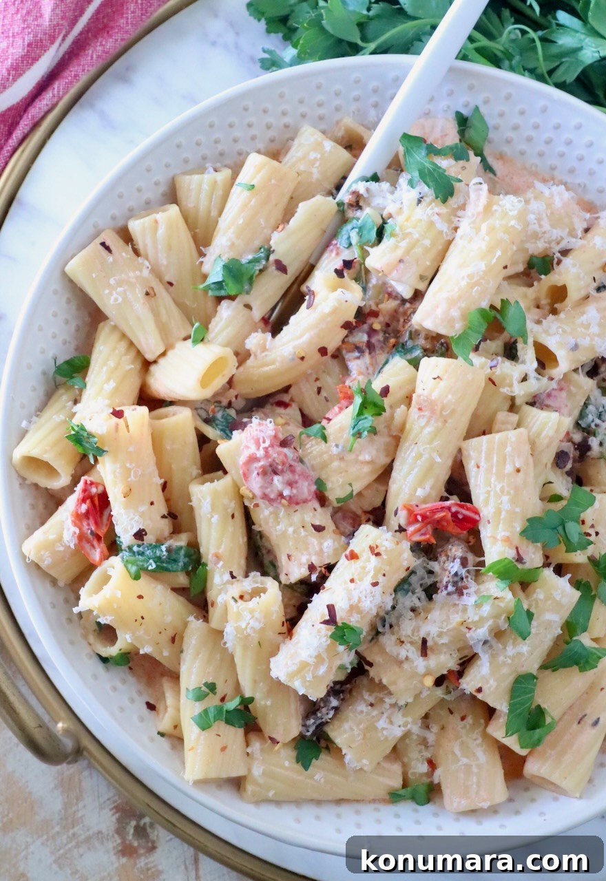 A bowl of creamy rigatoni pasta topped with fresh parsley, grated cheese, and red pepper flakes, mixed with sun-dried tomatoes; a fork rests in the bowl.