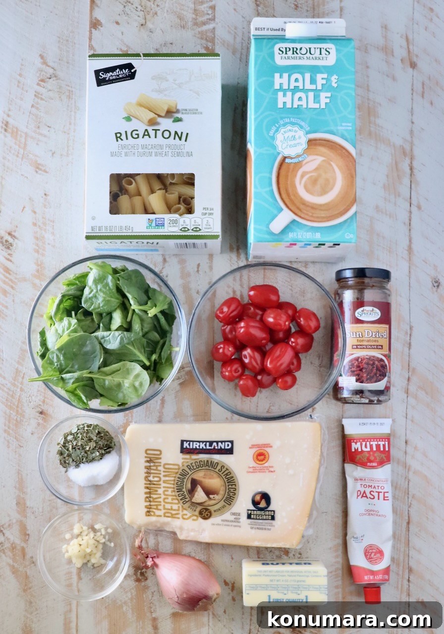 Assorted ingredients on a light wooden surface, including rigatoni, half & half, spinach, cherry tomatoes, garlic, herbs, parmesan, tomato paste, shallot, butter, and sun-dried tomatoes.