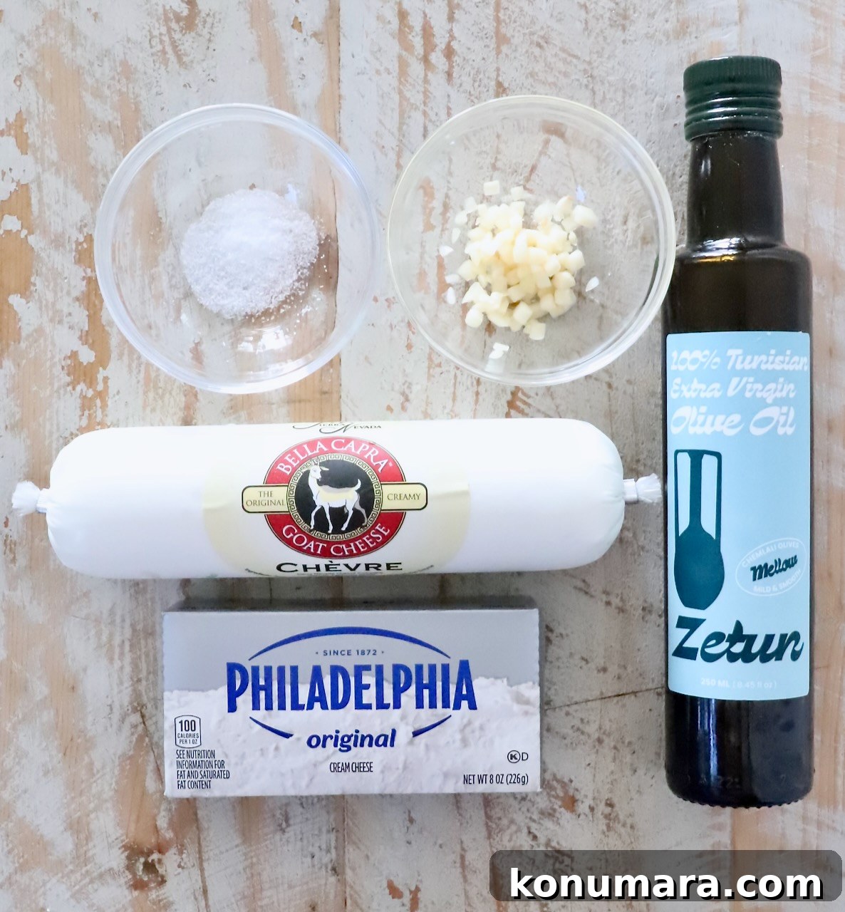 ingredients for goat cheese dip on a white wood board including olive oil, cream cheese, goat cheese, minced garlic and salt