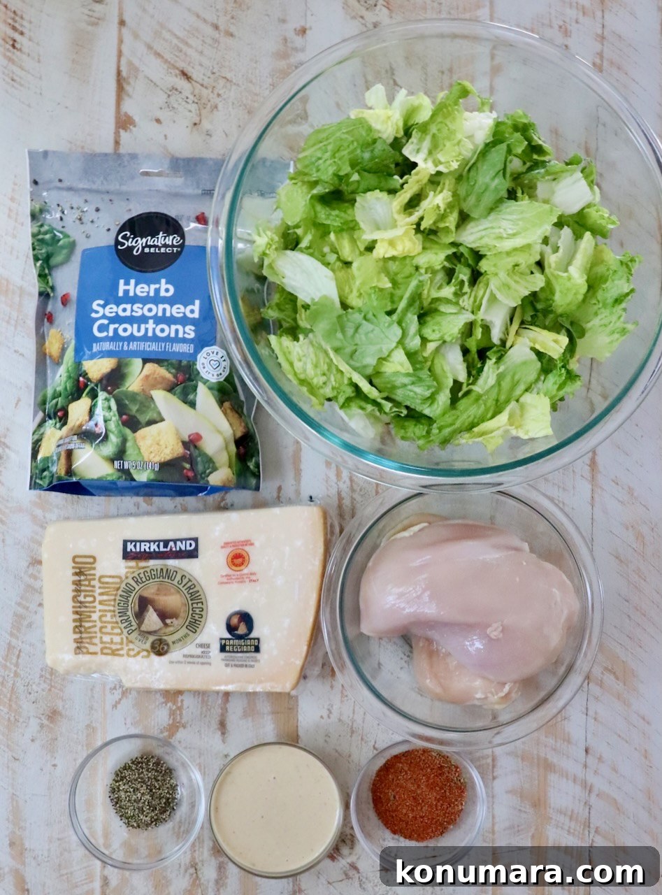 ingredients on a white wood board include a bowl of chopped romaine lettuce, a bag of croutons, parmesan cheese, raw chicken breasts, creamy dressing, and spices