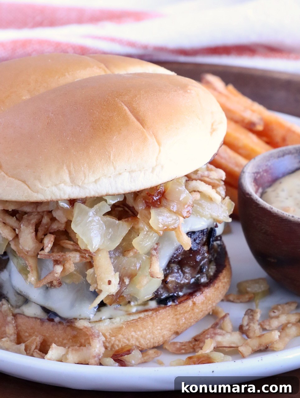 cheeseburger on a plate topped with crispy onions, with a side bowl of sauce, and sweet potato fries behind the burger on the plate