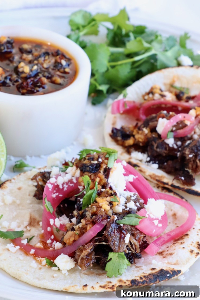 two tacos on a plate topped with smoked beef cheek barbacoa, pickled onions, salsa, cotija cheese and cilantro
