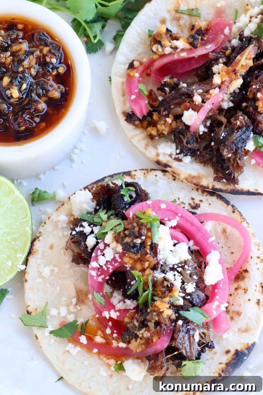 smoked beef cheek tacos on a plate with salsa macha, fresh cilantro, pickled onions and a lime wedge
