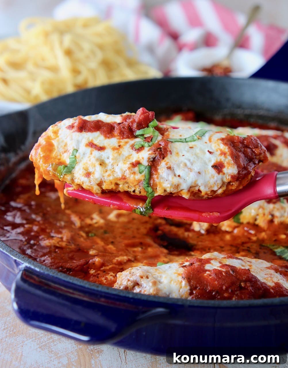 Red spatula holding a piece of chicken parmesan out of a large blue skillet