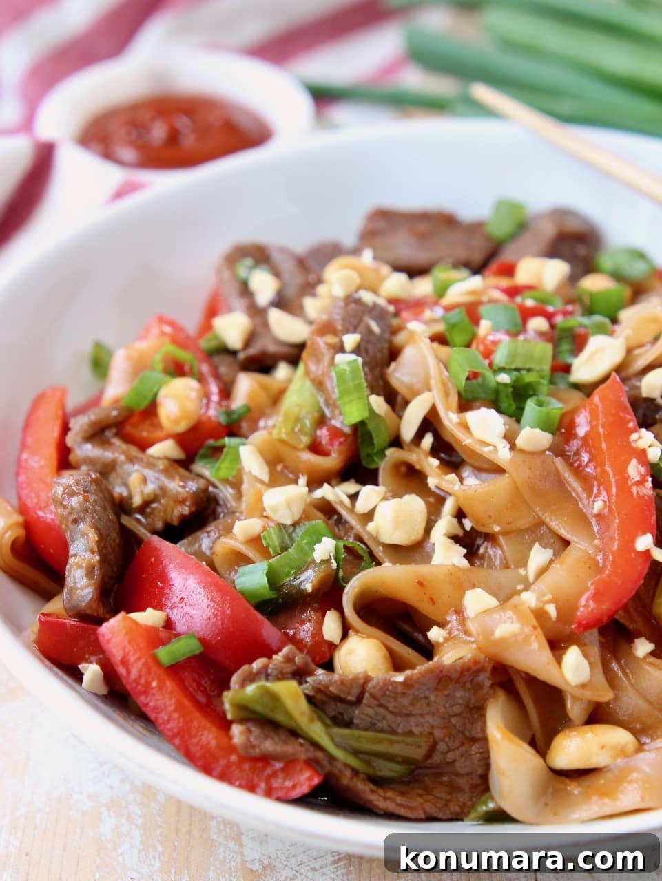 Kung pao beef, rice noodles and red bell peppers in white bowl topped with peanuts and green onions