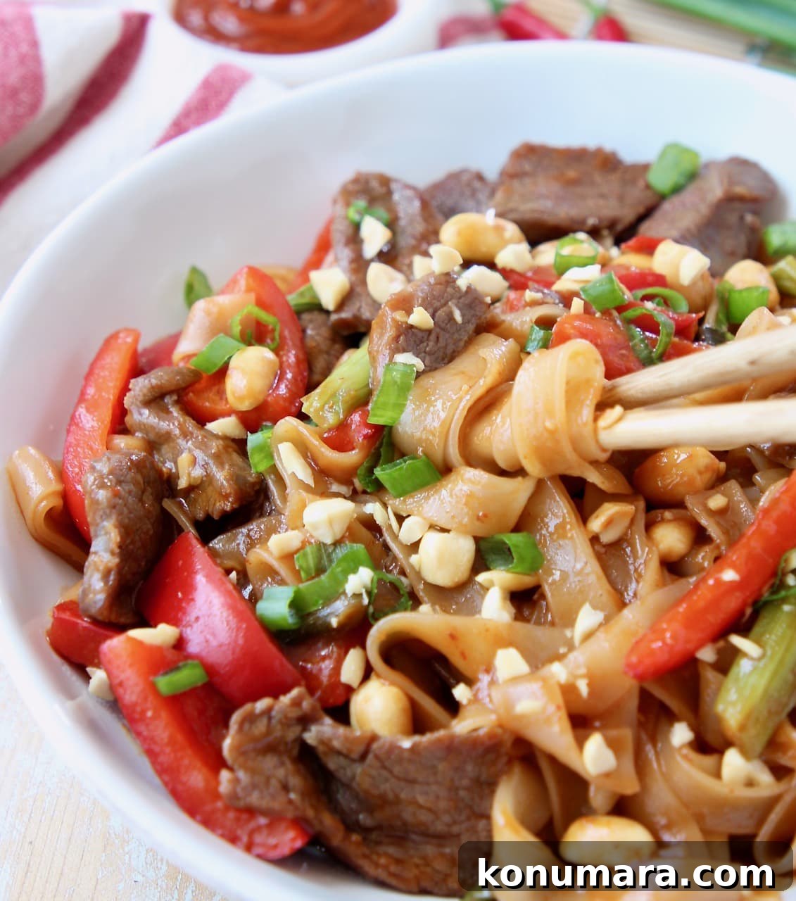 Noodles wrapped around chopsticks in bowl with beef, bell peppers and peanuts