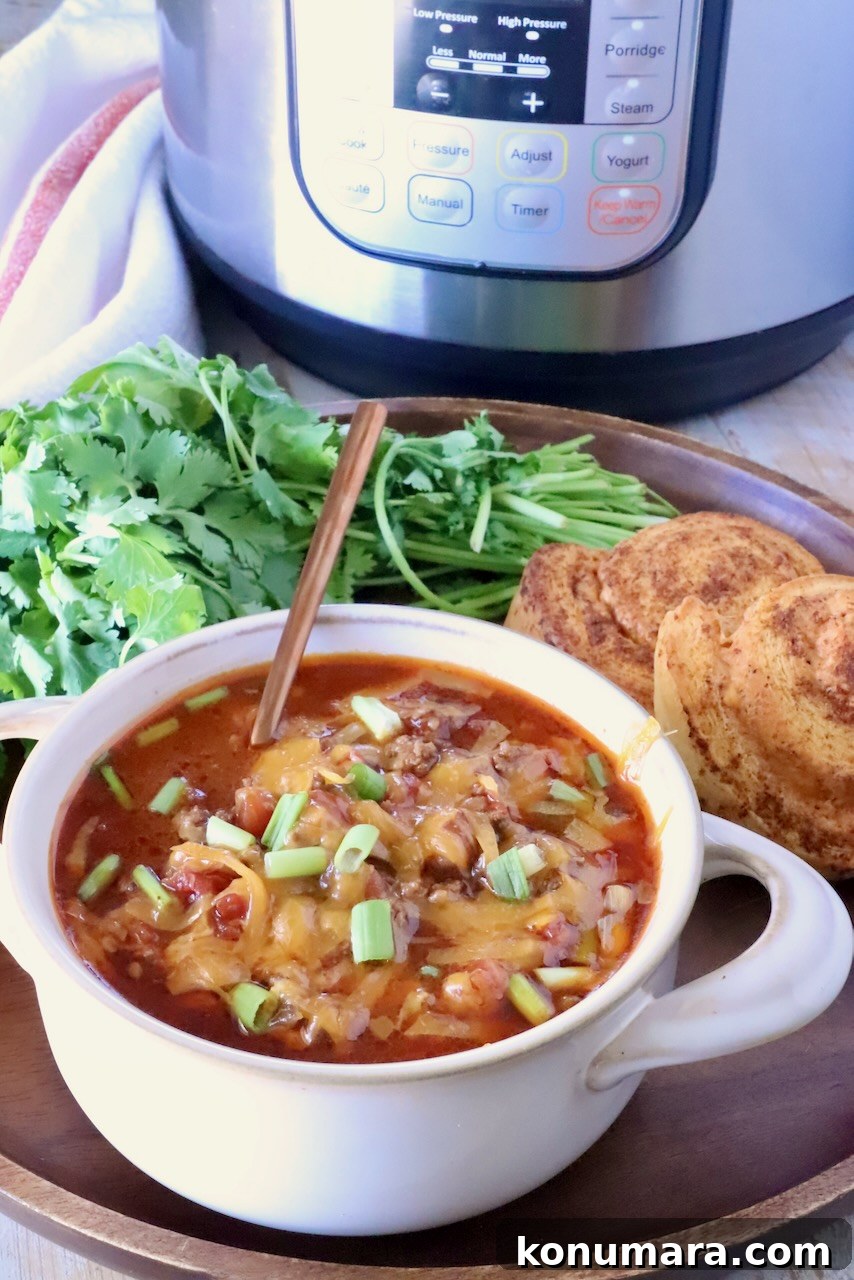 chili in a bowl with an Instant Pot, fresh cilantro, and cornbread rolls behind it