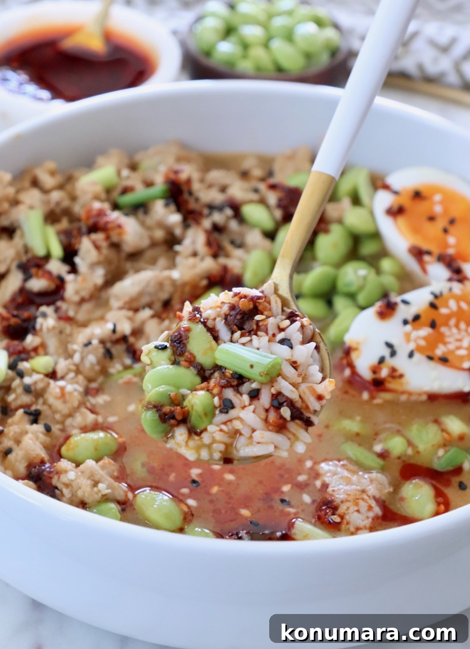 spoon scooping rice and edamame from a broth bowl with ground chicken, a jammy egg, and chili crisp on top